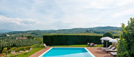 A rectangular swimming pool with a clear blue water surface, surrounded by a brick deck. There are lounge chairs and umbrellas on the deck, and a neatly trimmed hedge to the right. In the background, there are rolling hills and a clear sky.