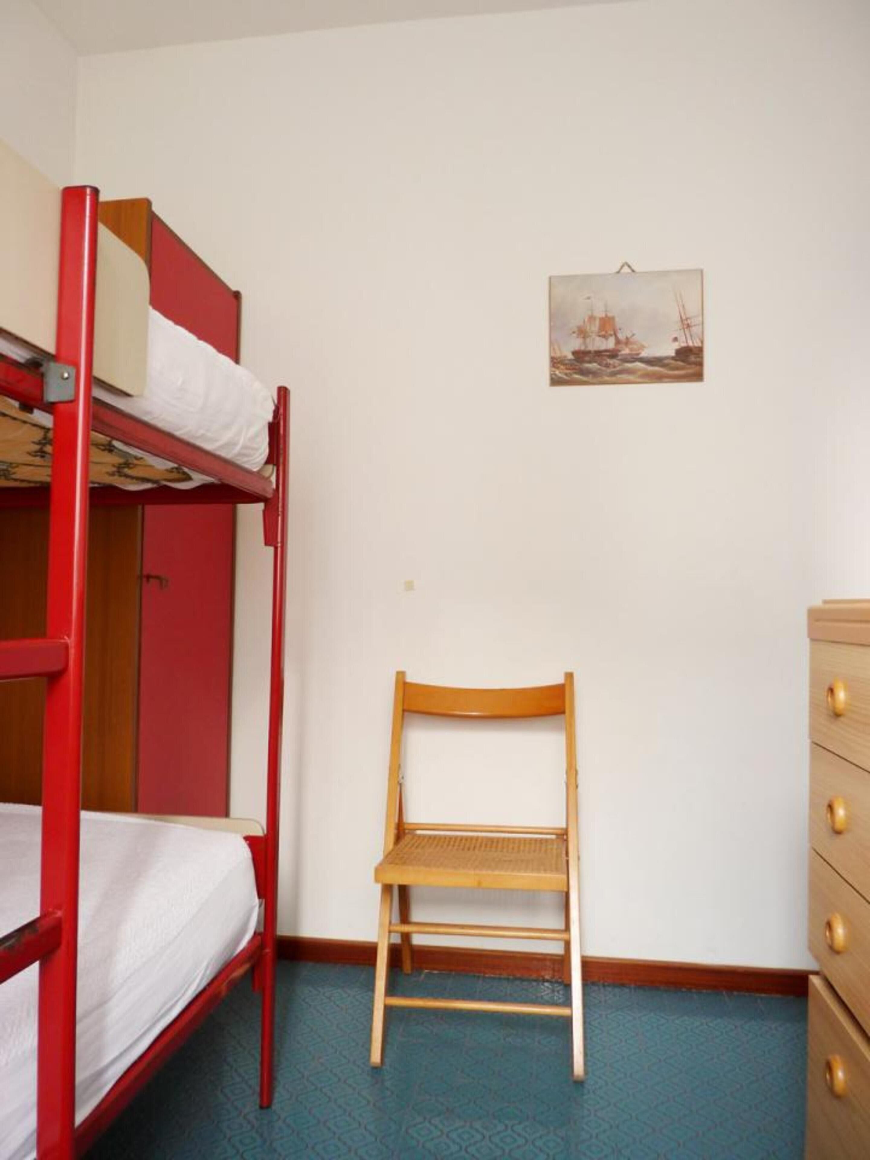Un lit superposé avec une structure rouge et une chaise en bois, dans une pièce dont le sol est recouvert d’un motif bleu.