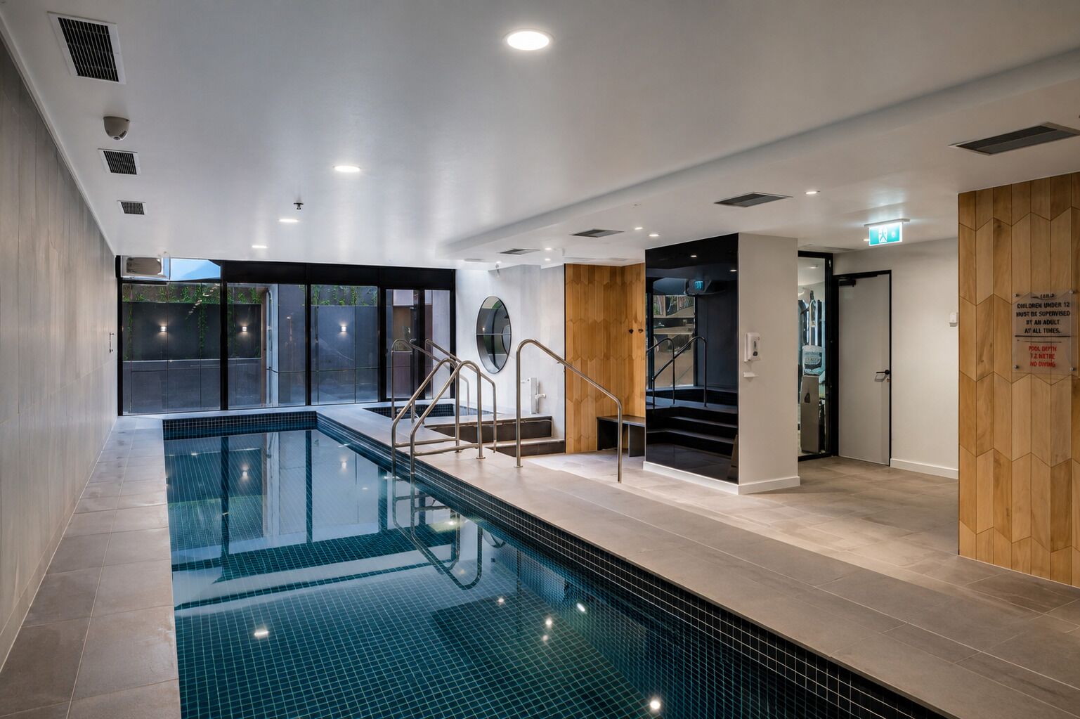 A modern indoor swimming pool with clear glass walls, a wooden wall panel, and a staircase leading to a lower level.