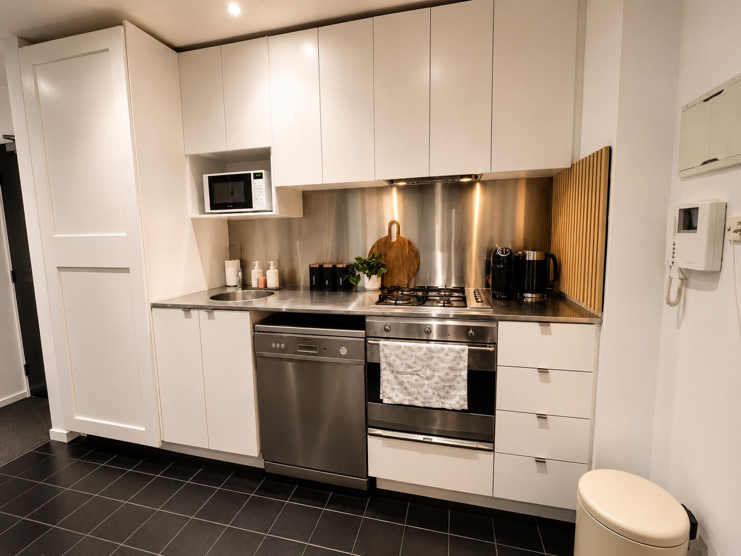 Une cuisine moderne dotée d’appareils électroménagers en acier inoxydable, de meubles blancs et d’un sol carrelé noir.