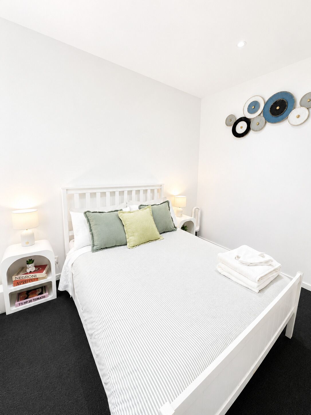 A white bed with a striped bedspread, two pillows, a bedside table with a lamp, a small shelf with books, and a wall decoration.