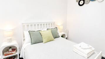 A white bed with a striped bedspread, two pillows, a bedside table with a lamp, a small shelf with books, and a wall decoration.