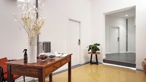 Un intérieur moderne avec une table en bois, un vase contenant des fleurs séchées, une petite plante en pot et une vue sur un couloir menant à une porte.