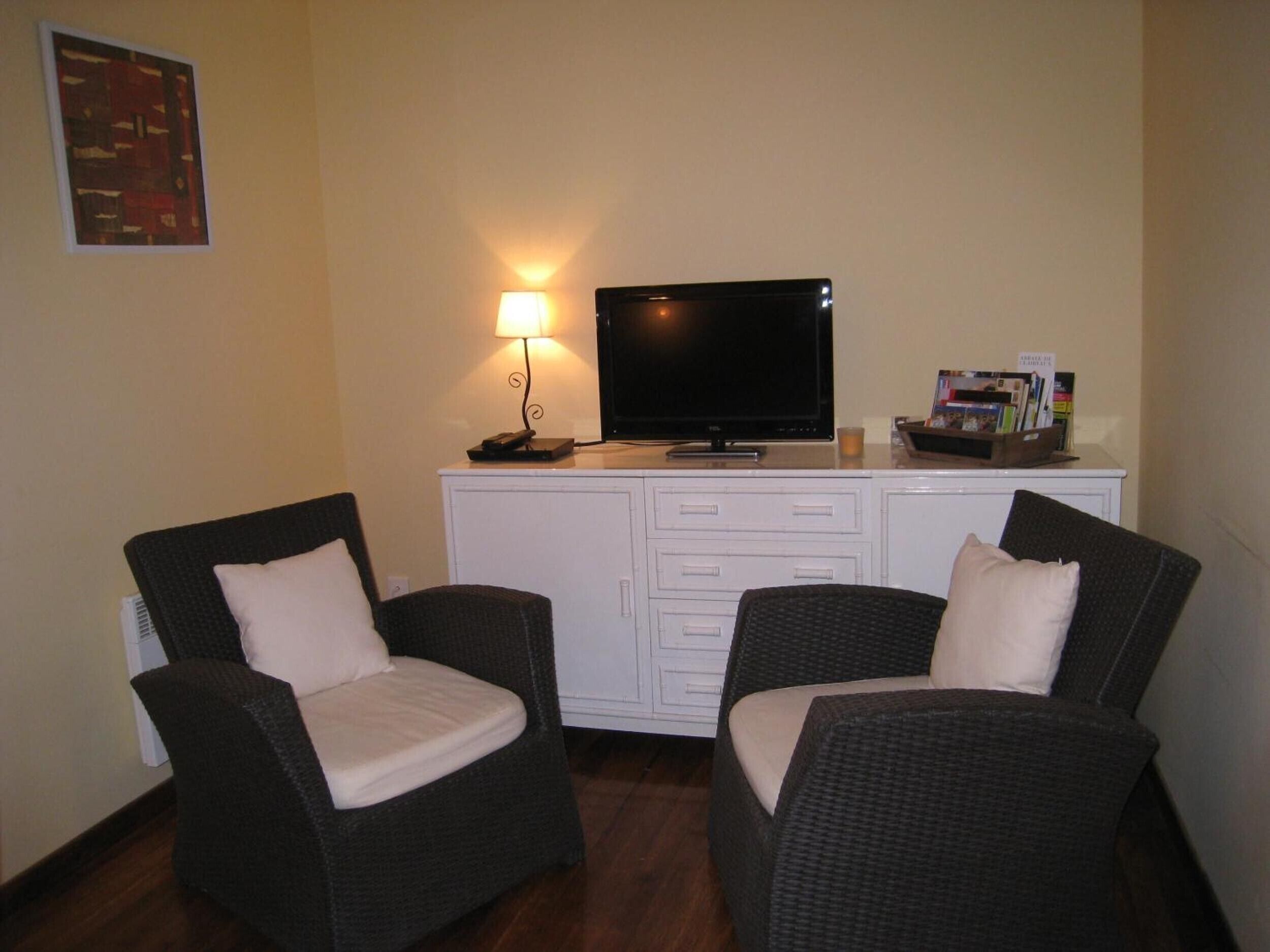 A room with two wicker chairs, a flat-screen TV, a desk with drawers, a lamp, and a framed picture on the wall.