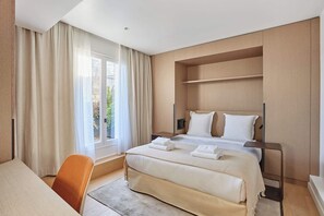 Habitación de hotel con una cama grande, un escritorio y una silla. Tiene una ventana con cortinas, y el piso y las paredes son de madera.