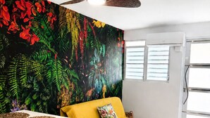 Ein Zimmer mit einem gelben Sofa, einem Esstisch, einem Deckenventilator und Wandbild.