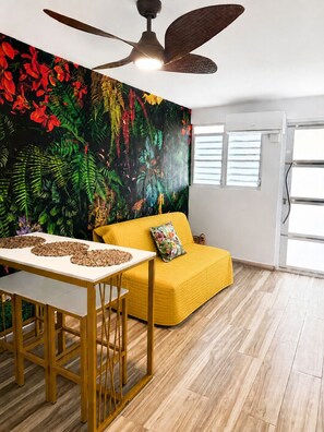 Une pièce avec un canapé jaune, une table à manger, un ventilateur de plafond et une fresque murale.