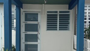 Ein Gebäudeeingang mit steinbelagtem Boden, einer Glastür mit Metallgriff und einem kleinen Fenster mit horizontalen Rollläden.