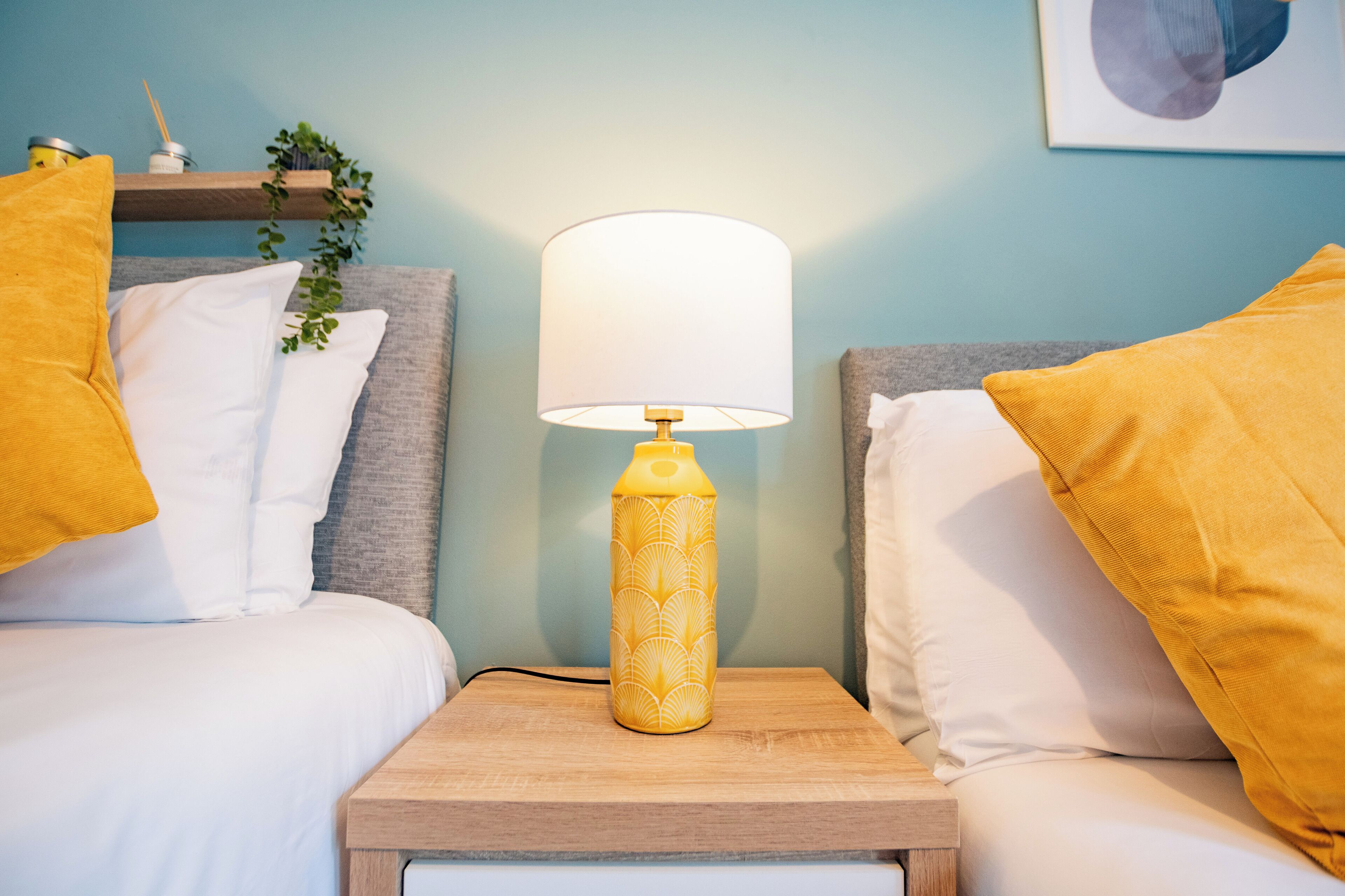 Un lit bien fait, avec une literie blanche et deux coussins jaunes, une table de chevet avec une lampe et une étagère murale agrémentée d’une plante.
