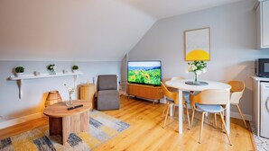 Ein modernes Wohnzimmer mit Flachbildfernseher, einem Esstisch mit Stühlen, einem Sofa, einem Couchtisch und einer Lampe.