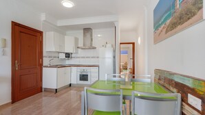 Eine Küche mit weißen Schränken, einer Mikrowelle und einem Kühlschrank. Ein Esstisch mit Stühlen steht bereit.
