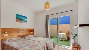 Ein Zimmer mit zwei Betten, einem Holzkopfteil, einem Bild, einem kleinen Tisch mit einer Pflanze und Blick auf eine grüne Außenanlage.