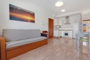 Uma sala de estar moderna com um sofá, cozinha com armários brancos e uma grande pintura de um pôr do sol.