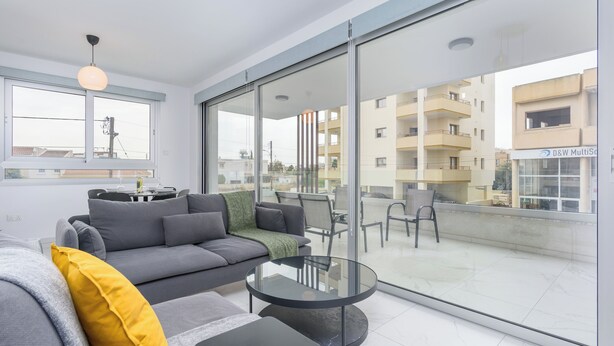 A modern living room with a grey sofa, a glass coffee table, and a balcony with a view of the city. - Mirage 2-BR Apt. & Balcony in Larnaca (Larnaca)