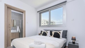 A bedroom with a bed, a nightstand, a window, and a bathroom door. - Mirage 2-BR Apt. & Balcony in Larnaca (Larnaca)