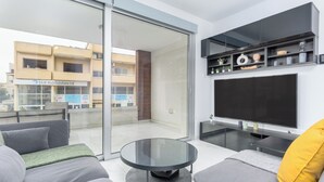 A modern living room with a glass sliding door leading to a balcony, a flat-screen TV mounted on the wall, and a round glass coffee table. - Mirage 2-BR Apt. & Balcony in Larnaca (Larnaca)