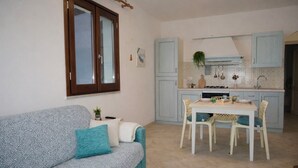 A compact living space with a sofa, a dining table with chairs, a kitchen area with cabinets and a stove, and a window with blinds. - Sea View Apartment with Terrace - Castelsardo | Montemarina (Castelsardo)