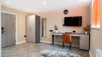 A modern room with a herringbone floor, a desk with a chair, a television mounted on the wall, and a wardrobe.