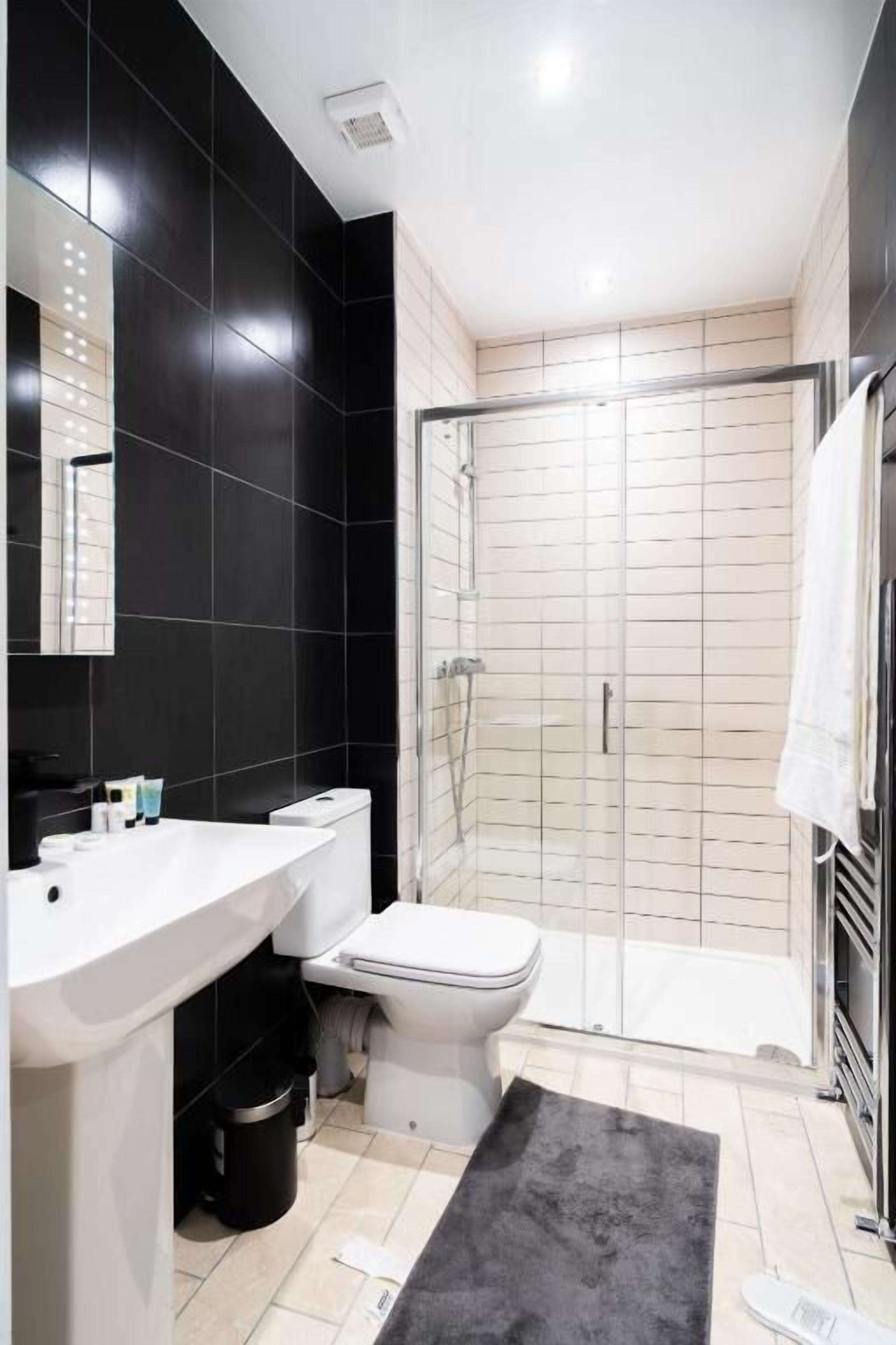 Un baño moderno con pared de azulejos negros, un lavabo blanco, un inodoro y ducha con mampara de vidrio.