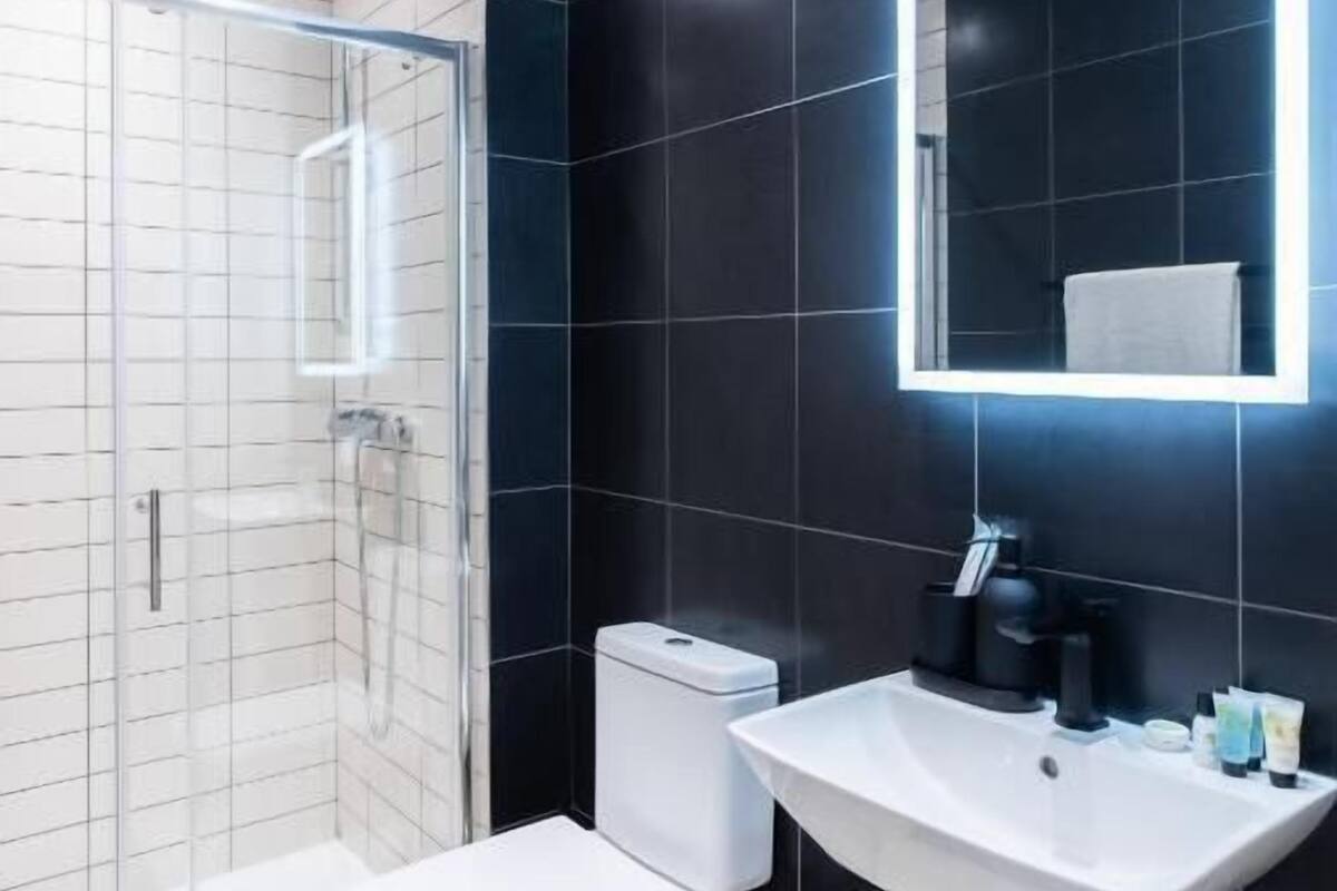 A modern bathroom with a glass shower enclosure, a white toilet, a pedestal sink, and a large mirror with underglow lighting.