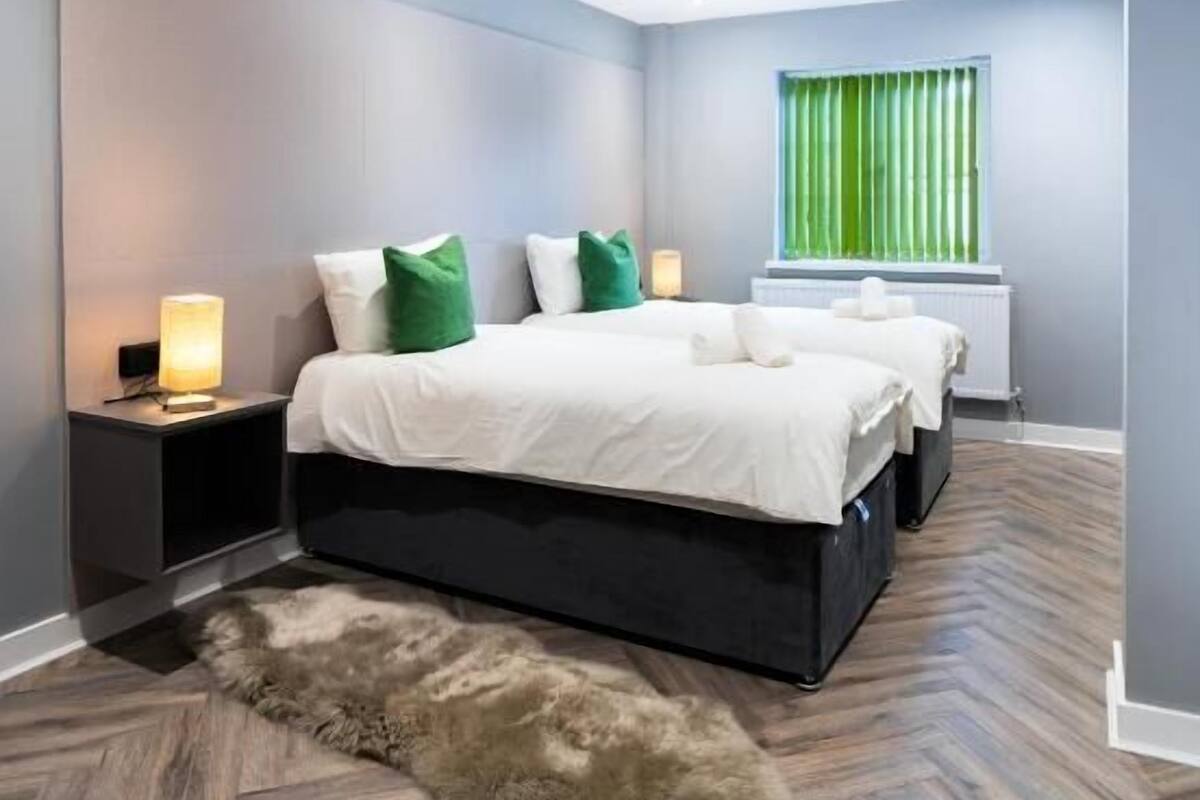 A modern bedroom with a large bed, two bedside tables, a fluffy rug, and a window with green curtains.