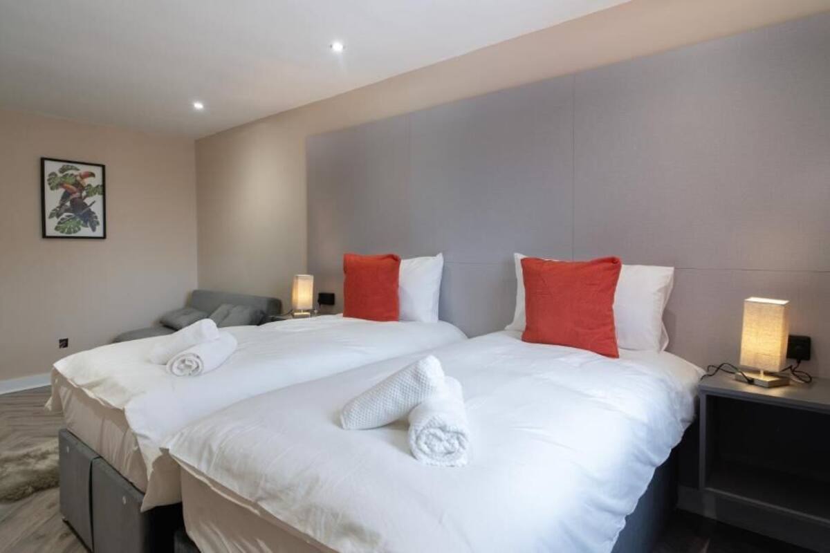 A hotel room with two beds, each with white bedding and red pillows. There are two bedside tables with lamps, a grey sofa, and a framed picture on the wall.