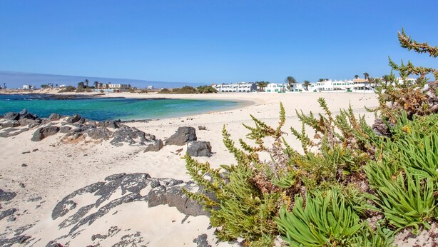 Ein Strand mit klarem, blauem Wasser, sandigem Ufer und Gebäuden im Hintergrund.