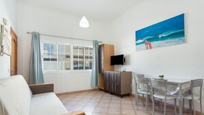 Ein Wohnzimmer mit einer Couch, einem Fernseher, einem Esstisch mit Stühlen, einem Schrank und einem Gemälde mit Strandmotiv.