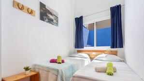 Ein Schlafzimmer mit zwei Betten, einem Fenster mit Vorhängen, einem Nachttisch mit Pflanze und einem an der Wand hängenden Bild.