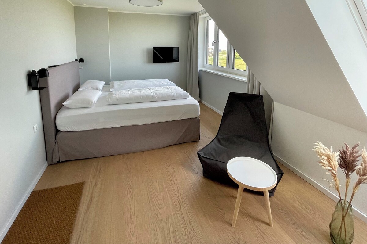 A modern bedroom with a bed, a flat-screen TV, a window, and a small table with a vase.