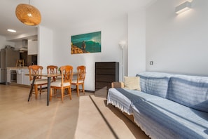 Een moderne woonkamer met een blauwe bank, een houten eettafel met stoelen, een schilderij aan de muur en een keuken op de achtergrond.