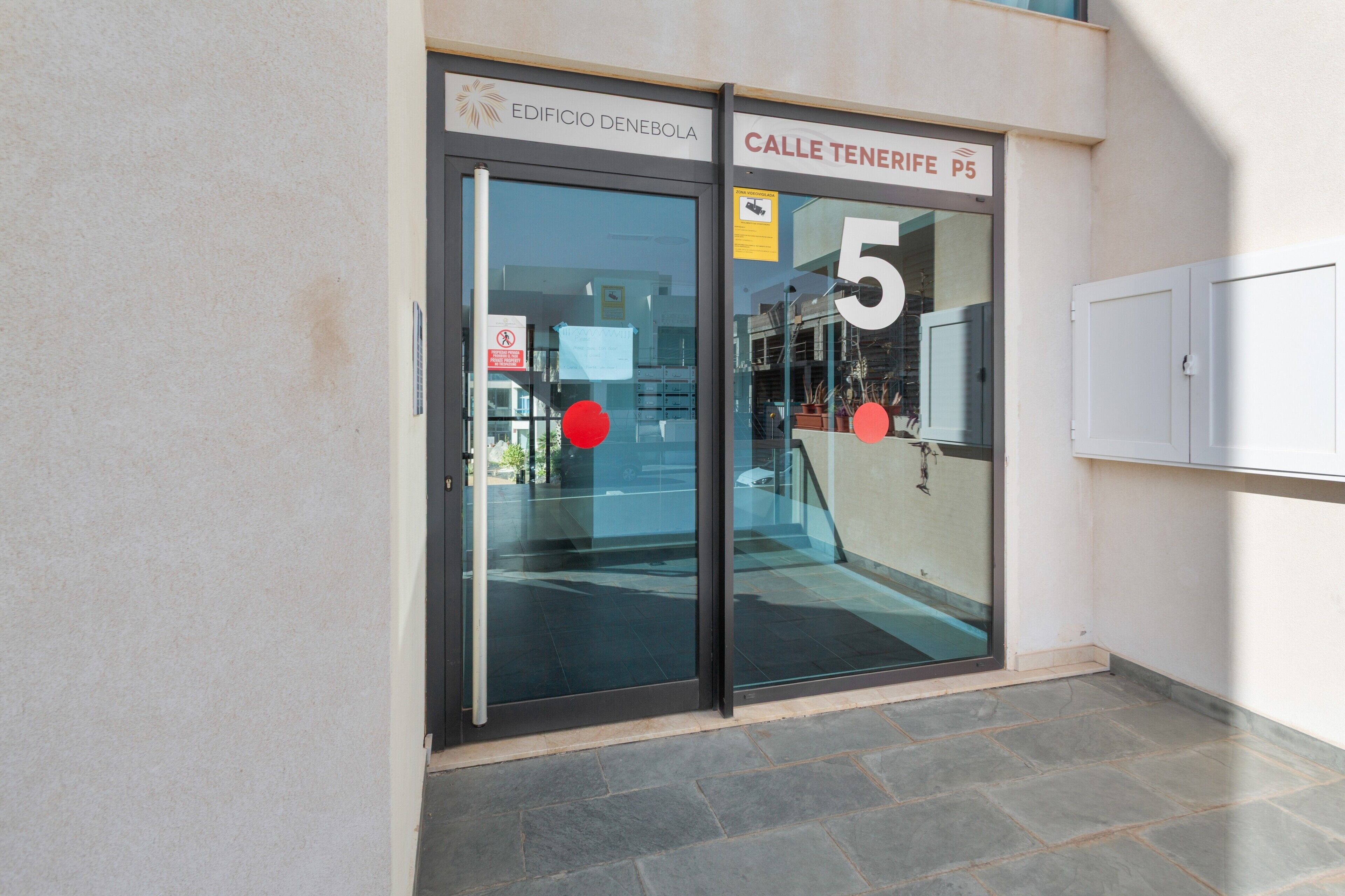 Entrada de un edificio con puerta de cristal, un cartel que dice 'EDIFICIO DENEBOLA' y una señal de calle que indica 'CALLE TENERIFE P5'.
