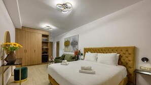 A modern hotel room with a large bed, a wooden wardrobe, a green armchair, and a small table with a vase of flowers. - Gold Suites (Ioannina)