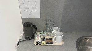 A kitchen counter with a coffee maker, a tray with coffee pods, milk, and sugar packets. A welcome sign with Wi-Fi and location details is visible on the wall. - 1066 Apart Hotel (Hastings)