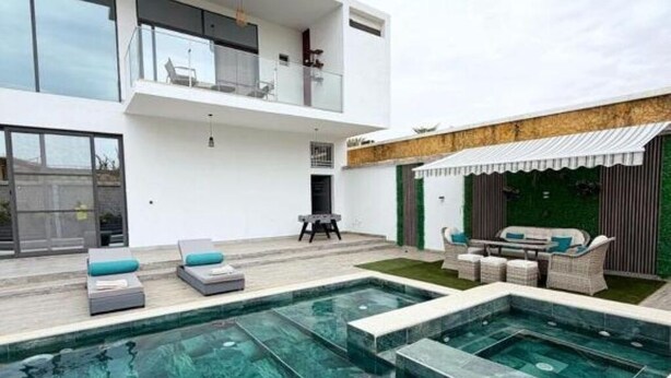 Un área de piscina moderna al aire libre con un edificio de dos pisos, mobiliario exterior y una zona de estar cubierta.