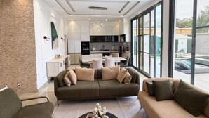 Una sala de estar moderna con un sofá y una mesa de centro, una zona de cocina con armarios blancos y ventanas amplias que ofrecen vistas al exterior.