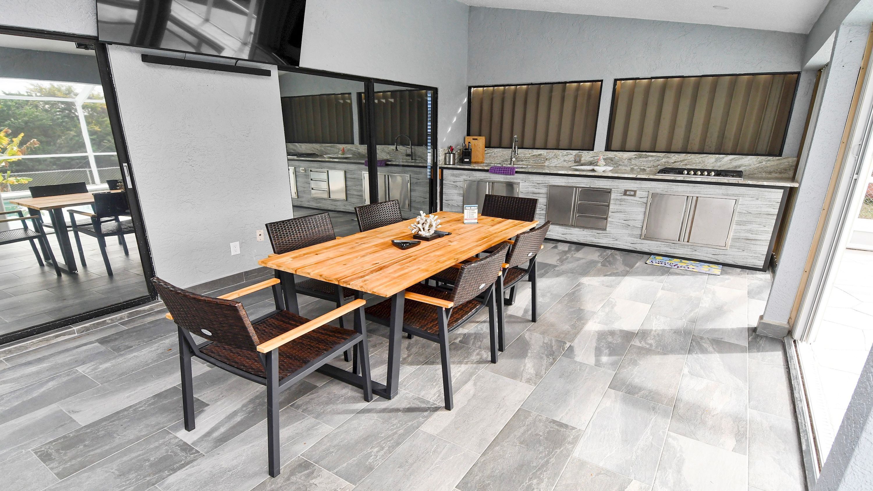 A modern kitchen with a marble countertop, a dining area with a wooden table and wicker chairs, and a large window with blinds.
