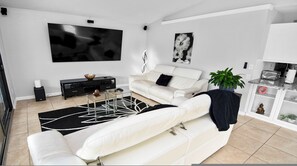 Un salon moderne avec un canapé blanc, une table basse en verre, un tapis noir et blanc, une console noire ornée d’un vase décoratif, un téléviseur fixé au mur et une plante en pot.
