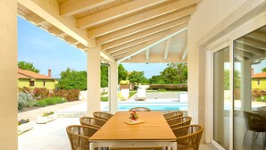 An outdoor covered patio with a wooden table and wicker chairs, overlooking a swimming pool and a residential area. - Villa Corsa by Villas Guide (Barban)
