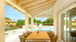 Eine überdachte Terrasse im Freien mit einem Holztisch und Korbstühlen, mit Blick auf einen Swimmingpool und eine Wohngegend.