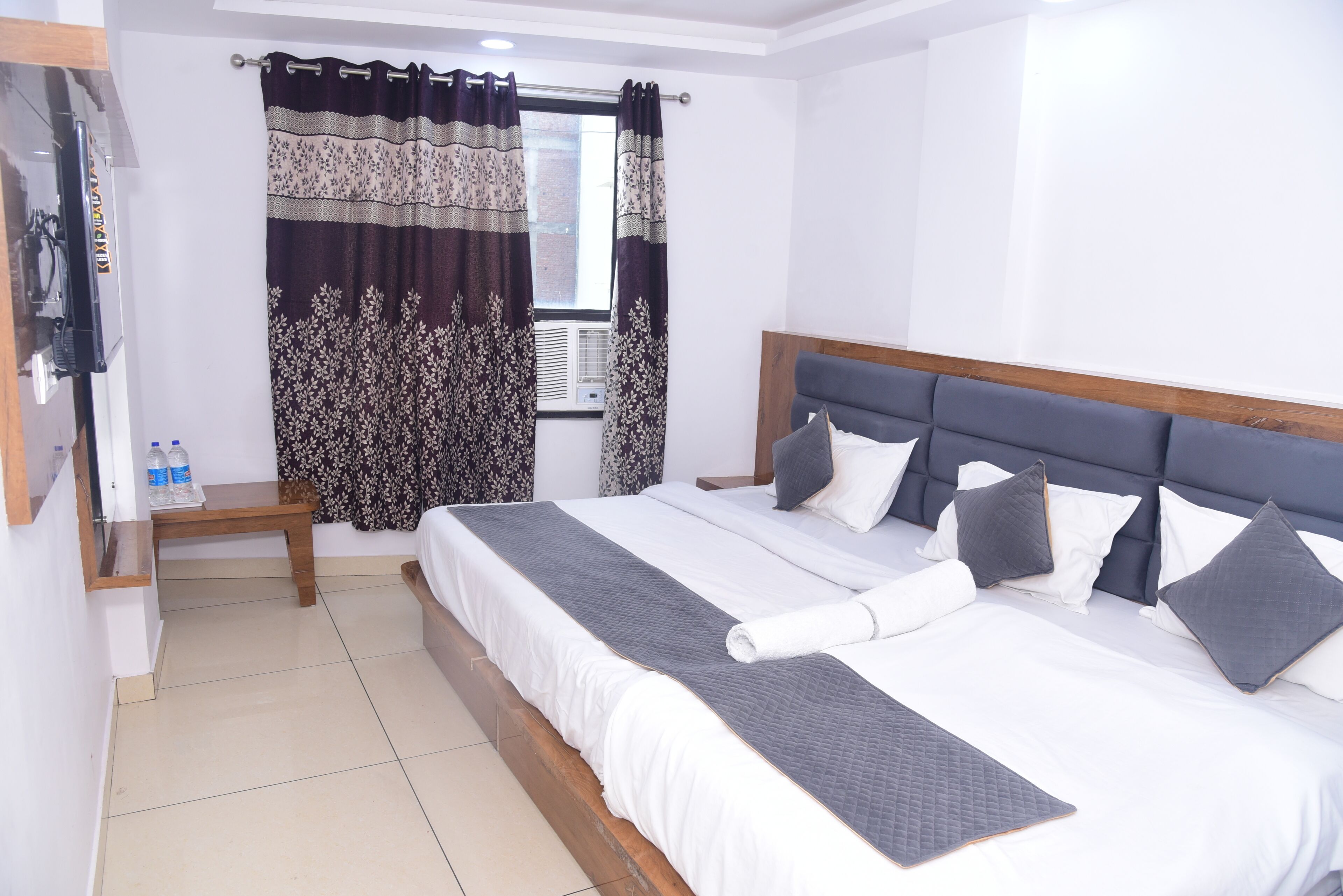 A bedroom with a bed, a bedside table, a window with curtains, and a wall-mounted TV.