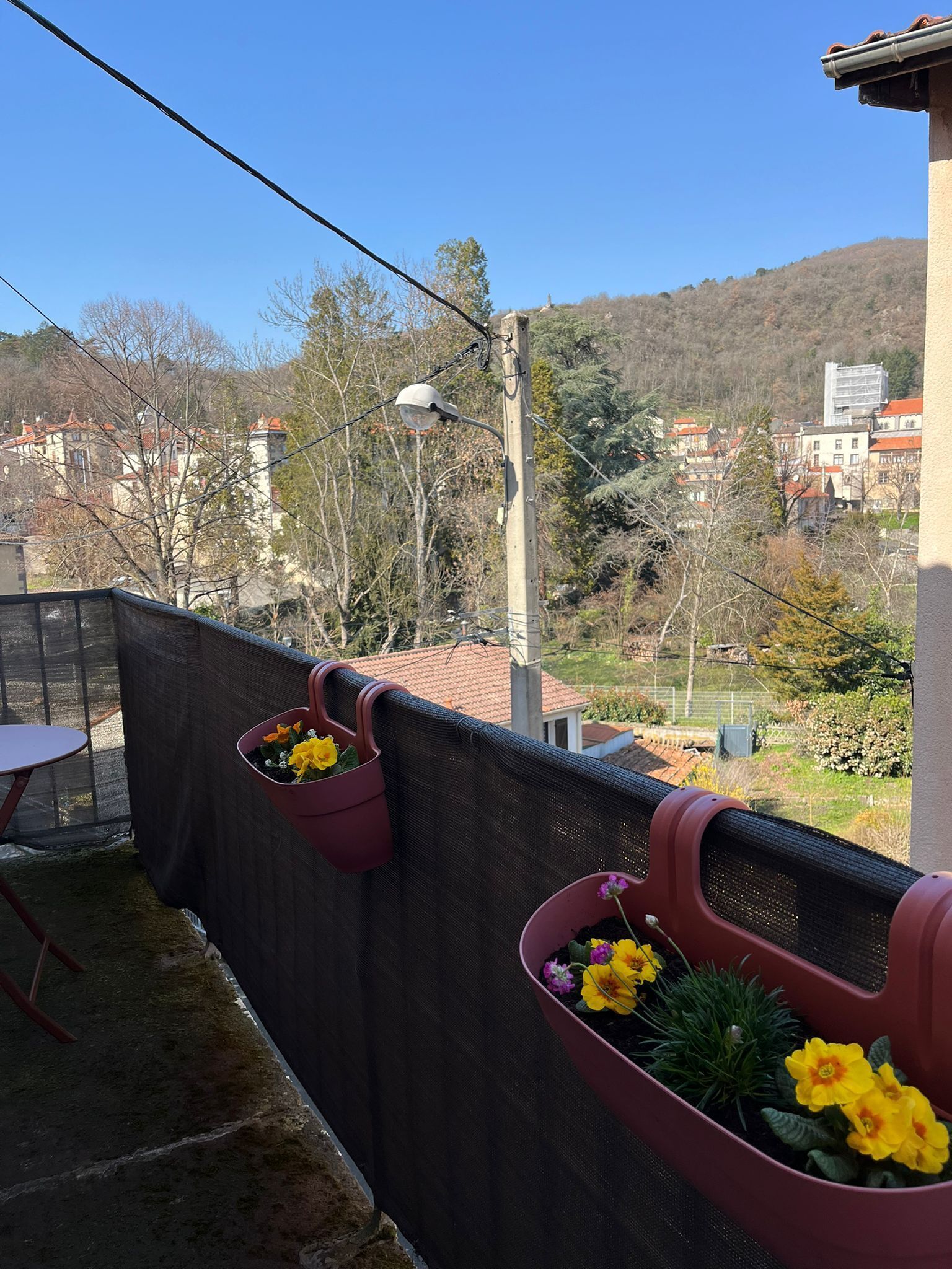 Un balcon avec deux jardinières contenant des fleurs jaunes, une table et une vue sur des bâtiments et des arbres en arrière-plan.