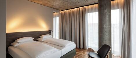 A modern hotel room with a large bed, a seating area, and a large window with curtains.