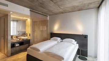 A modern hotel room with a large bed, a bedside table, a bathroom with a sink and mirror, and a view of the outdoors.