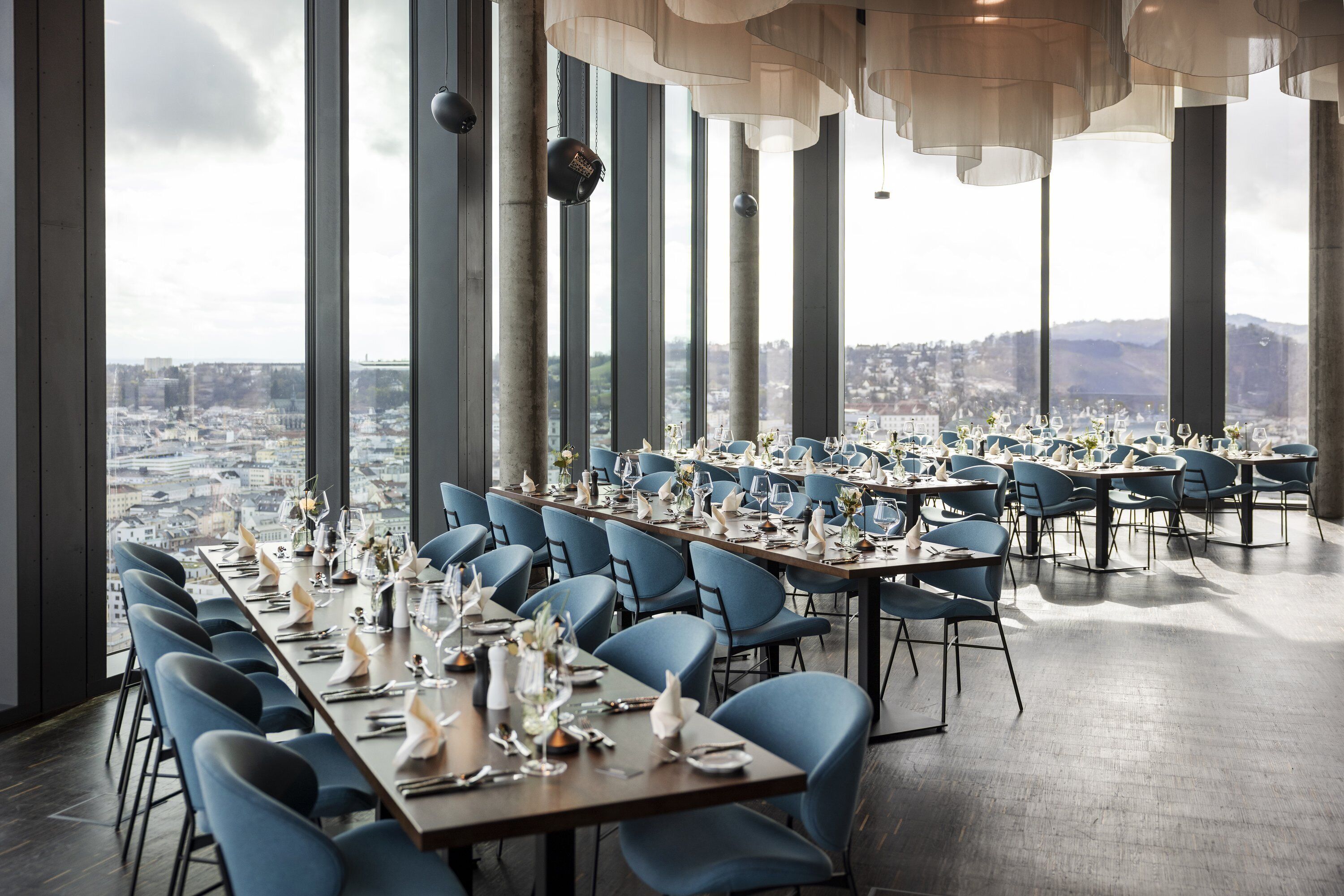 A restaurant with long tables, blue chairs, and a city view.
