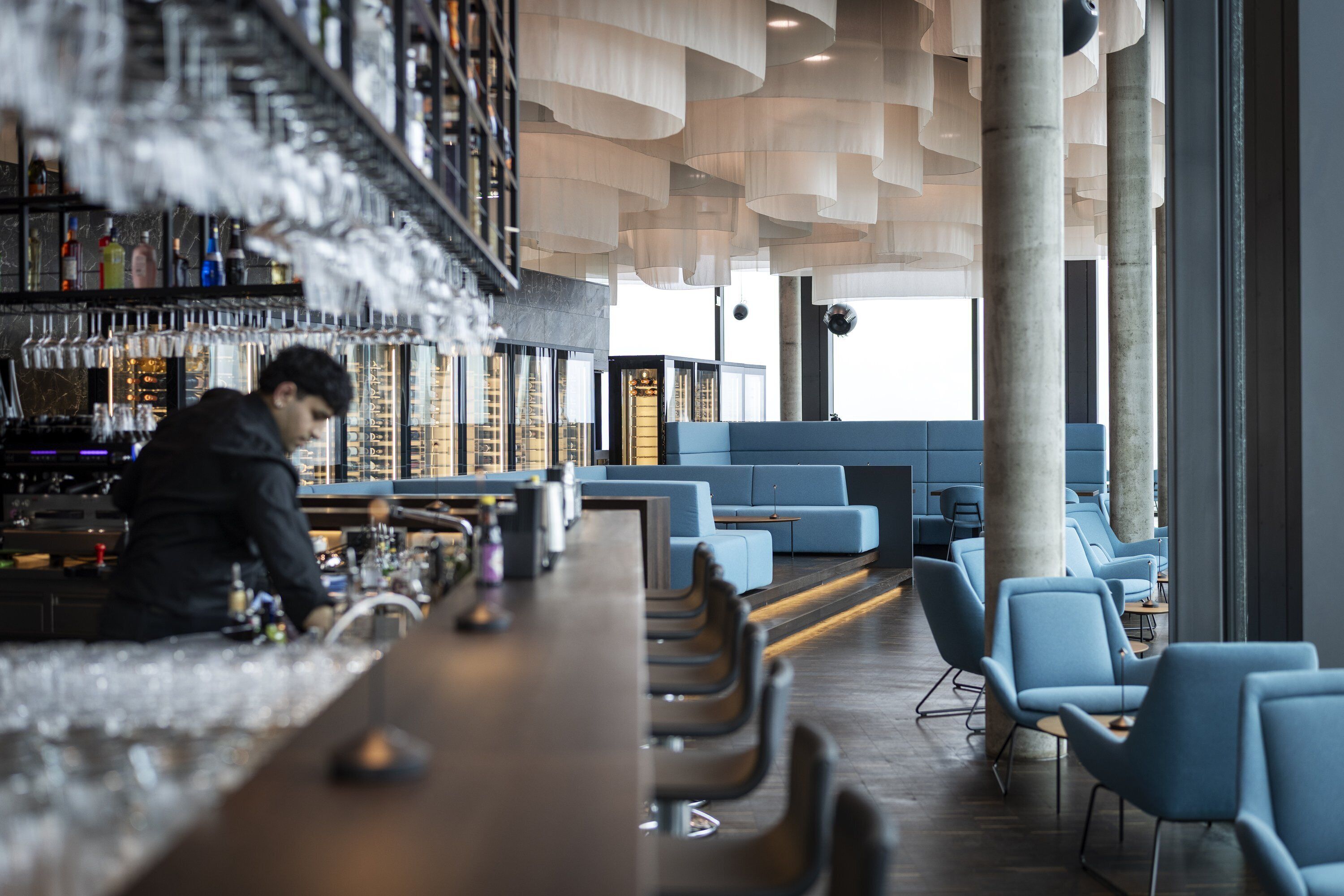 Eine Bar mit Barkeeper, Sitzgelegenheiten mit blauen Stühlen und einem modernen Deckenstil.