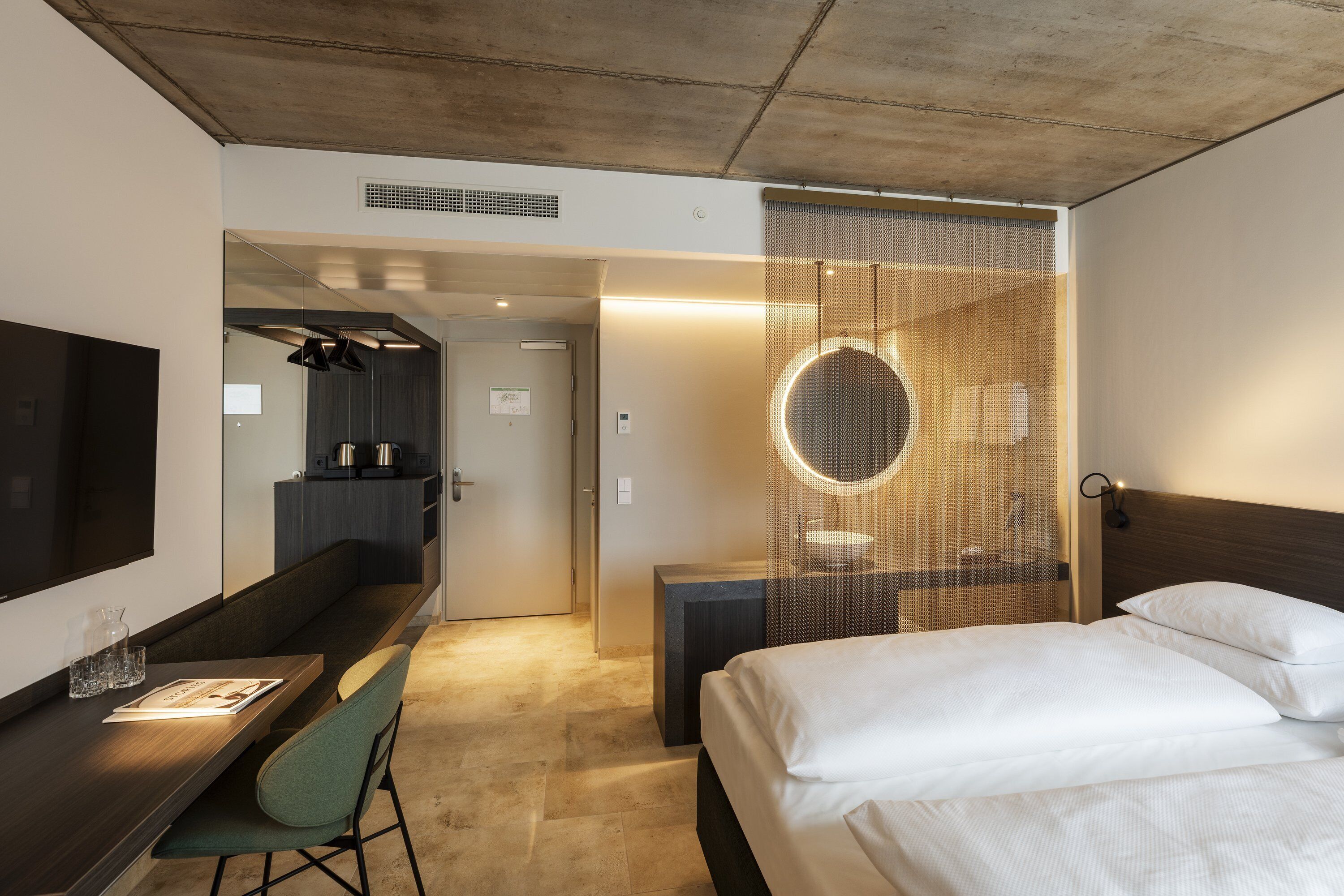 Ein modernes Hotelzimmer mit Bett, Schreibtisch, Stuhl, Fernseher und einem Badezimmer mit rundem Spiegel.