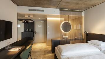 Ein modernes Hotelzimmer mit Bett, Schreibtisch, Stuhl, Fernseher und einem Badezimmer mit rundem Spiegel.