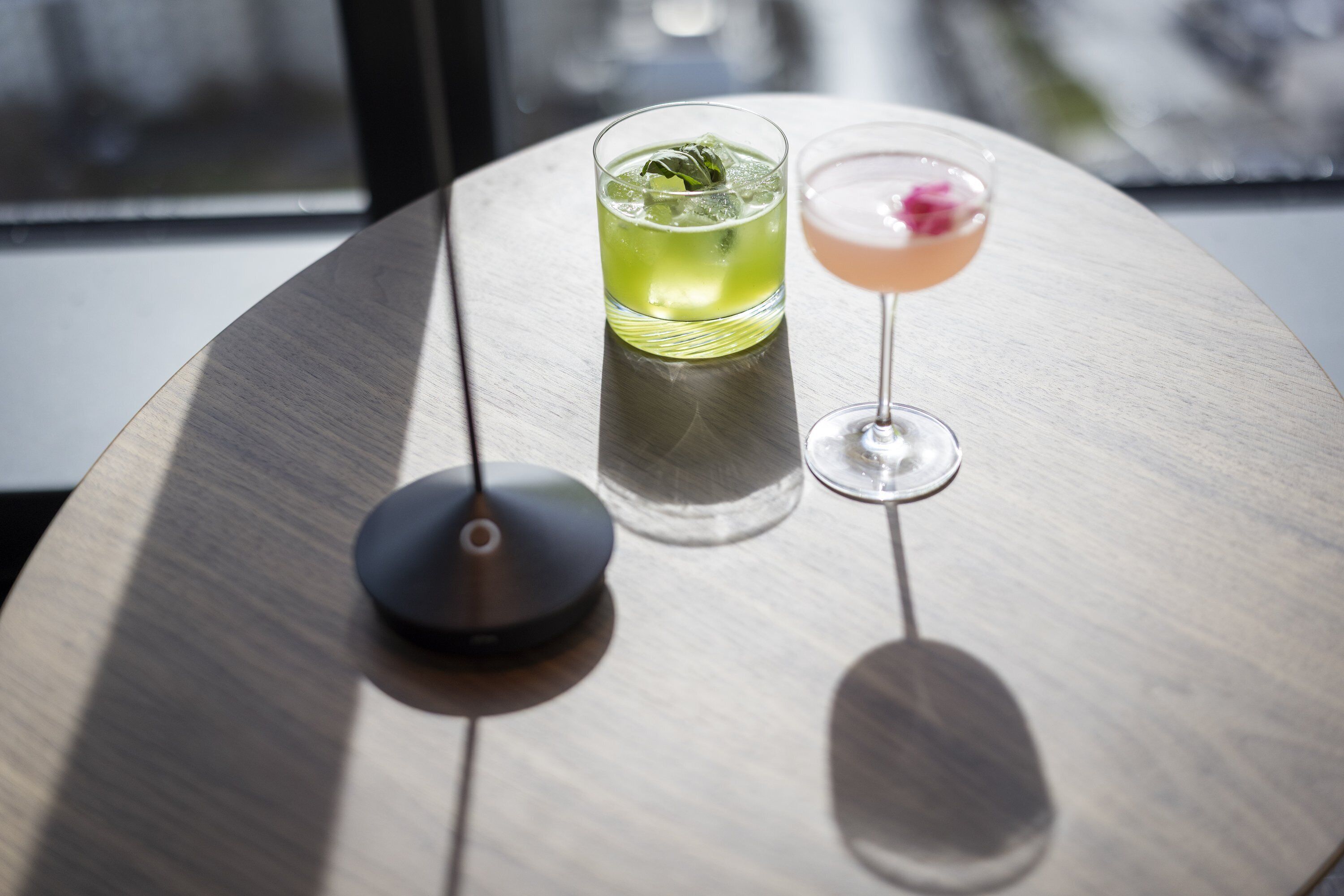 Dos vasos de bebidas sobre una mesa redonda de diseño moderno, un objeto negro pequeño y una ventana al fondo.