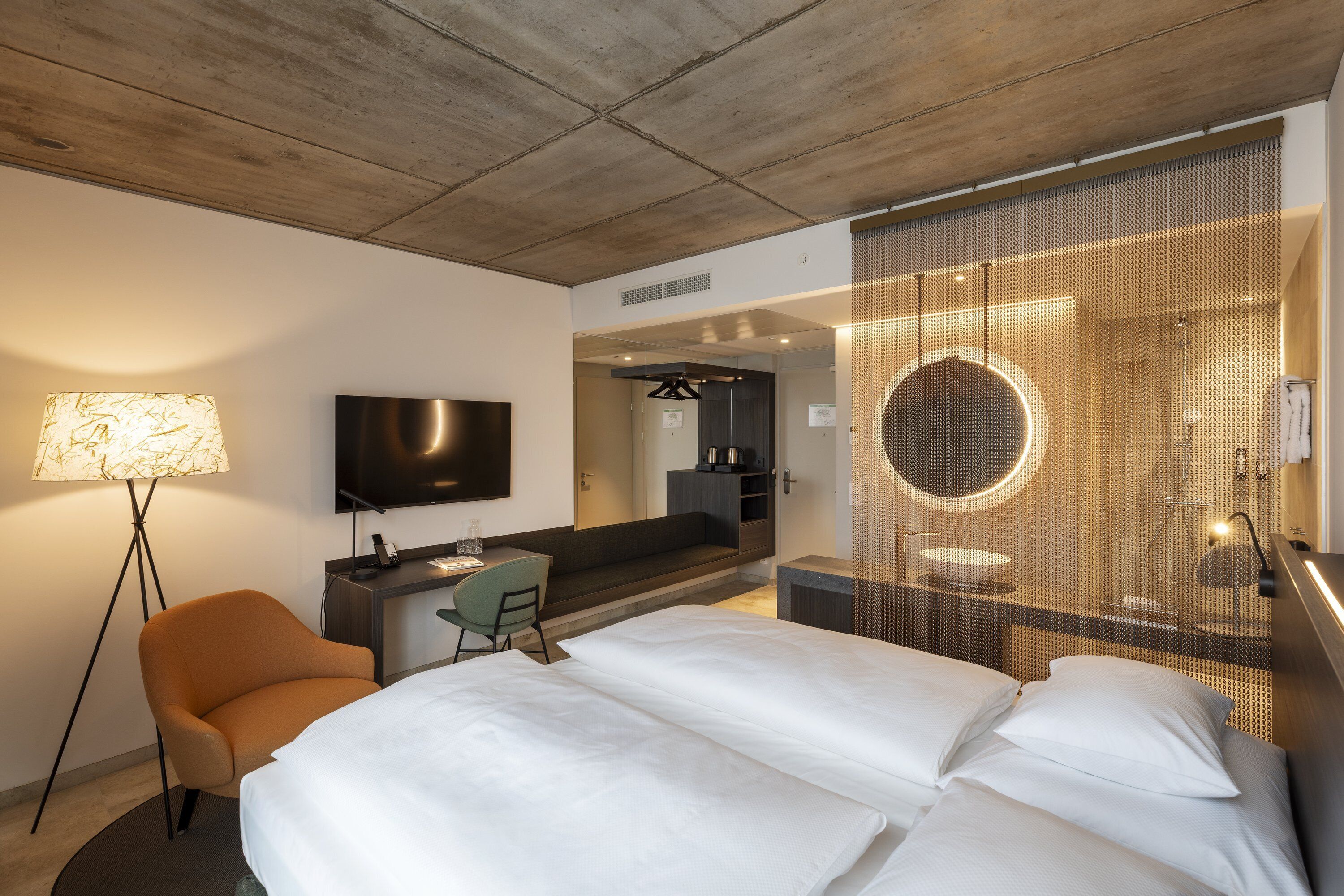 A modern hotel room with a large bed, a desk, a chair, a TV, and a shower area with a circular mirror.